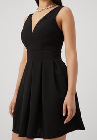 Černé šaty bez rukávů s výstřihem do V, áčkovou plisé sukní a přiléhavým korzetem. Hladká textura a minimalistické designové akcenty.