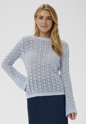 Frau trägt einen hellblauen Spitzenstrickpullover und einen dunklen Rock, steht mit einer Hand in der Hüfte vor einfachem Hintergrund.