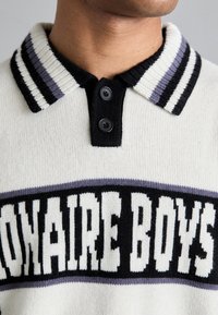 Kremine megztinėje polo su juodais ir purpuriniais dryžuotais raišteliniais kraštais, su dviem juodais mygtukais ir dideliais užrašais "Billionaire Boys".