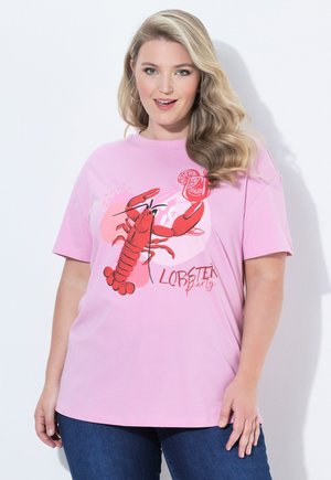 Hymyilevä nainen, jolla on laineikkaat vaaleat hiukset, pukeutunut vaaleanpunaiseen T-paitaan, jossa on punainen hummeri cocktailin kanssa ja teksti "Lobster Party", yhdistettynä sinisiin farkkuihin.