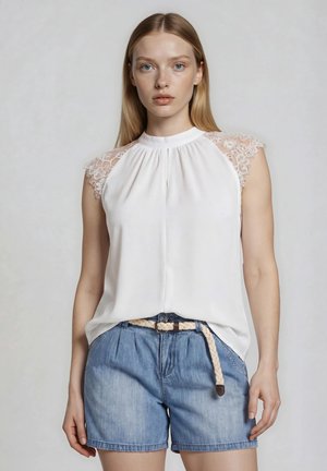 Jeune femme portant un chemisier blanc sans manches avec des épaules en dentelle et un short en jean bleu avec une ceinture tressée, debout devant un fond uni.