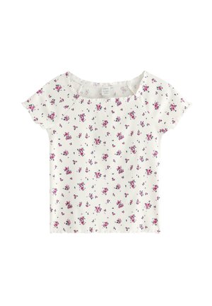 Pige iført en lilla kasket, hvid blomsterkortærmet top, mørkeblå jeans og beige sneakers står mod en ensfarvet hvid baggrund.