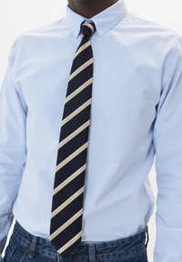 Corbata navy con rayas diagonales beige, de tela texturizada, llevada sobre una camisa de botones azul claro. Jeans de mezclilla visibles en la cintura.