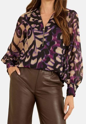 Vrouw die een bruine leren broek draagt en een blouse met lange mouwen met een abstract patroon in paars, zwart en beige en een rits aan de voorkant.