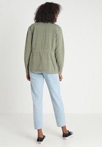 Olive green, lightweight jacket with a slightly oversized fit, worn over light blue jeans and black slip-on shoes, viewed from the back.