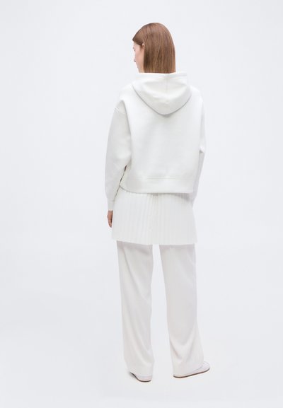 Femme aux cheveux bruns raides portant un sweat à capuche blanc surdimensionné, une jupe plissée, un pantalon ample et des chaussures blanches, vue de dos sur un fond blanc.