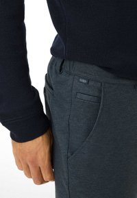 Pantaloni strutturati grigio scuro con frontale piatto, tasche laterali e una piccola etichetta del marchio, abbinati a un maglione lavorato a maglia blu navy.