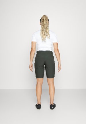 Persona de espaldas, vestida con una camiseta blanca de manga corta, pantalones cortos oscuros hasta la rodilla, zapatos negros y con el cabello largo y rubio trenzado.