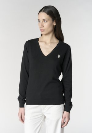 Donna con lunghi capelli scuri che indossa un maglione nero con scollo a V e un piccolo logo ricamato, e pantaloni bianchi, che guarda di lato.