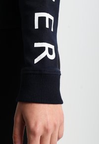 Maglietta a maniche lunghe blu navy con testo bianco sulla manica. Presenta polsini a costine e una texture liscia.