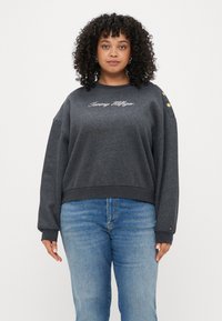 Grå sweatshirt med rund halsringning, med broderad text "Tommy Hilfiger" och gyllene knappar på axeln. Långa ärmar med ribbad mudd.