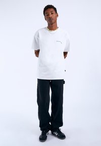 Bílý tričko s krátkým rukávem a malou zelenou grafikou, v kombinaci s volnými černými kalhotami a černými sportovními botami. Jako doplněk je použita korálková náhrdelník.