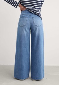 Osoba w szerokich niebieskich dżinsach i topie w marynarskie biało-niebieskie paski z długim rękawem, stojąca z jedną ręką na biodrze.