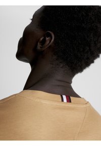 Närbild av en person med kort lockigt hår som bär en beige topp med en randig etikett i rött, vitt och svart vid bakhalsen.