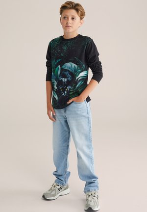 Maglietta nera a maniche lunghe con un design giungla verde che presenta una pantera, abbinata a jeans azzurri chiari e sneakers grigie con accenti neri.