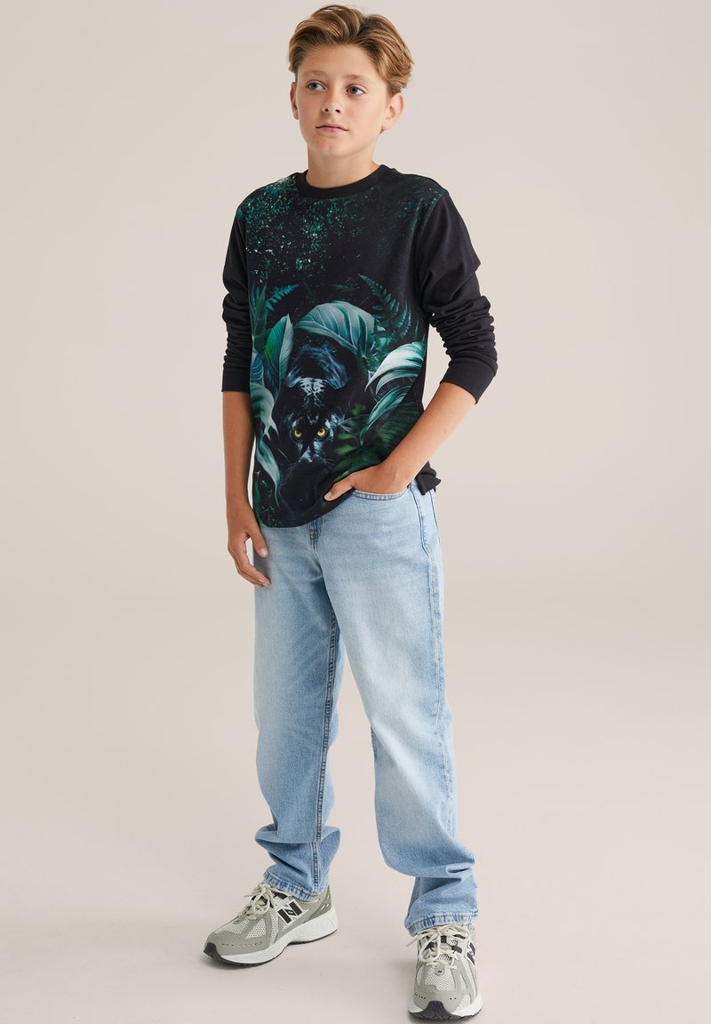 Mustassa pitkähihkaisessa paidassa on vihreä viidakkokuosi, jossa on pantteri, ja se on yhdistetty vaaleansinihin farkkuihin sekä harmaisiin tennareihin, joissa on mustia yksityiskohtia.