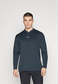 Svart langermet skjorte med halv glidelås og krage, laget av glatt stoff, med et logo på brystet og rett hem i bunnen.
