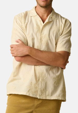 Mann verschränkt die Arme, trägt ein kurzärmeliges, hellgelbes, strukturiertes Blumenhemd und senffarbene Hosen vor einem einfarbigen Hintergrund.