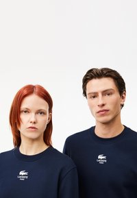 Felpe blu navy con scollo rotondo, caratterizzate da ricamo del logo Lacoste bianco sul petto, indossate da due modelli.
