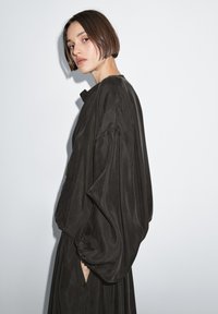 Schwarzes, fließendes Kleidungsstück mit weiten Ärmeln und hohem Kragen. Glatte Textur mit gerafften Stoffdetails, die elegant fällt.
