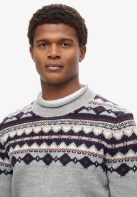 Grijze gebreide trui met patroonaccenten in navy, crème en bordeaux. Heeft een hoge kraag en een textuurafwerking.