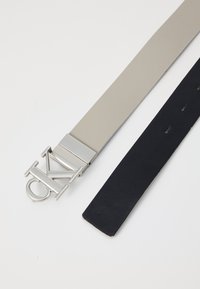 Twee riemen: één in glad lichtgrijs materiaal met een metalen logo-sluittang, en een andere in gestructureerd donkerblauw leer met perforaties.