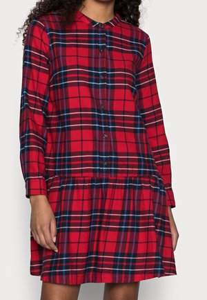 Frau trägt ein rotes kariertes langärmliges Kleid mit Knöpfen vorne und einem gerafften Rock, steht vor einem einfarbigen Hintergrund.