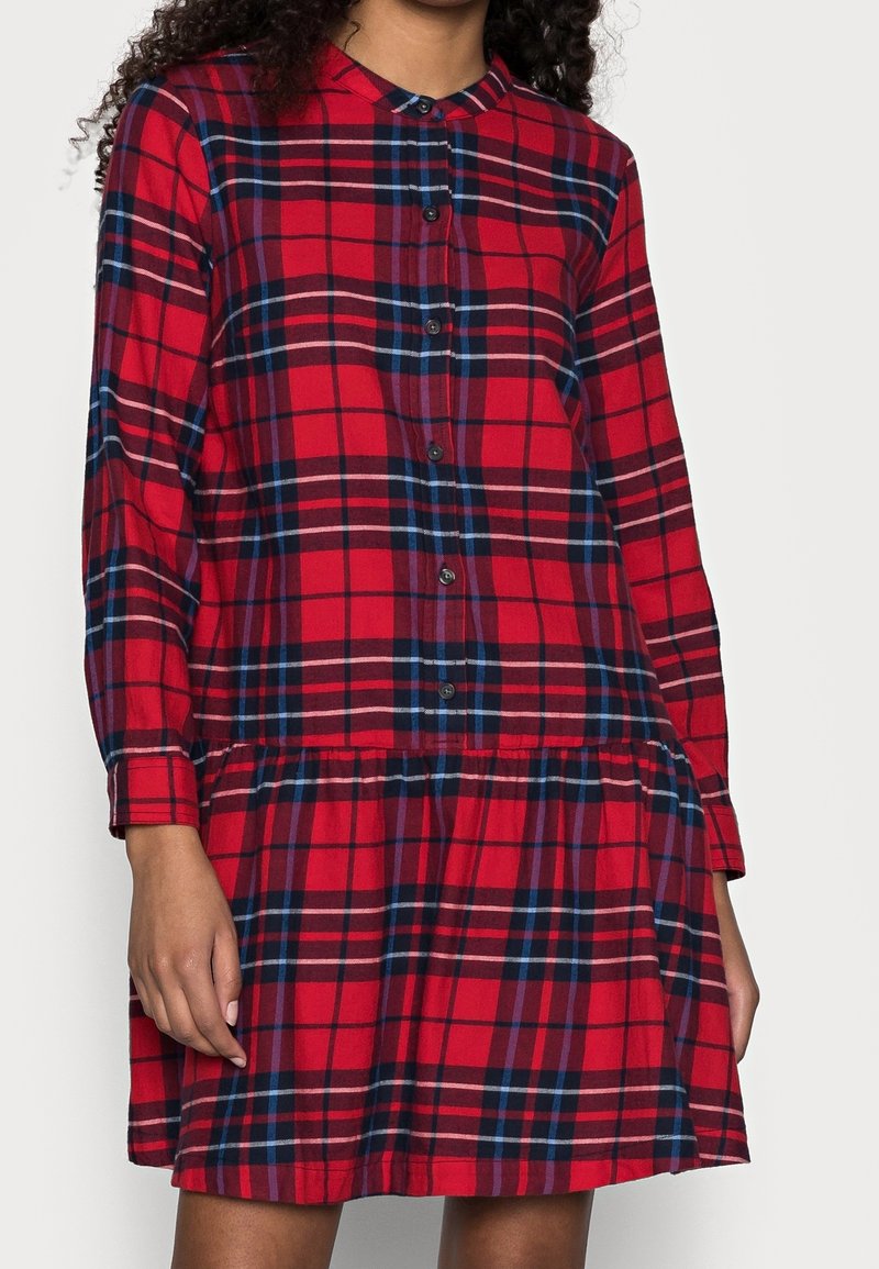 Femme portant une robe à carreaux rouges à manches longues avec des boutons sur le devant et une jupe froncée, debout devant un fond uni.