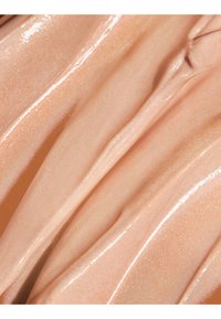 Crème ou lotion beige scintillante avec une texture lisse et brillante, étalée en couches épaisses et ondulées.