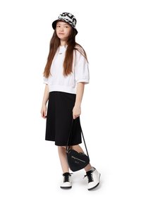 Pull blanc court avec une légère brillance, jupe noire jusqu'aux genoux, sac à main noir en forme de cœur, et baskets noir et blanc avec des lacets.