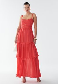 Robe maxi en chiffon rouge à fines bretelles, design superposé, accents à volants et corsage ajusté. Sac à main blanc et talons à fines lanières.