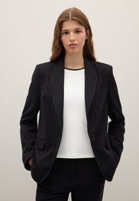 Blazer nero sartoriale con risvolti, chiusura a un bottone e due tasche frontali, indossato sopra una maglia bianca. Texture semplice, design strutturato.