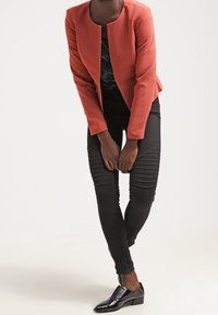 Veste orange rouille à encolure ronde, design ajusté et texture lisse, associée à un pantalon noir avec des accents côtelés.