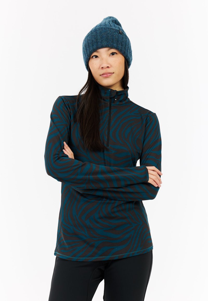 Camiseta de manga larga con cremallera, patrón de cebra en negro y verde azulado, tejido suave, cuello alto. Combinada con un gorro de punto azul.