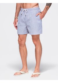 Homme portant un short rayé bleu clair et blanc avec cordon, des tongs beiges et des bracelets au poignet gauche, debout sur un fond uni.