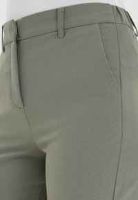 Nära bild av grågröna byxor med sidfickor, dragkedja och bälteshällor som bärs med en vit topp.