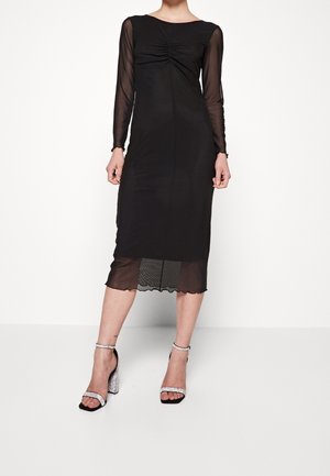 Kobieta nosząca czarną sukienkę midi z przezroczystymi długimi rękawami i dołem, w połączeniu ze srebrnymi sandałami na bloczku z brokatem.