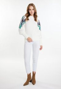 Witte fluffy trui met kleurrijke pailletten accenten op de mouwen, gecombineerd met lichtblauwe jeans en bruine suède enkellaarsjes.