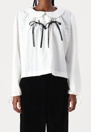 Henkilö, jolla on yllään valkoinen, pitkähihainen paita pyöreällä kaula-aukolla ja kaksi mustaa nauharusetia rinnassa, yhdistettynä mustiin housuihin.