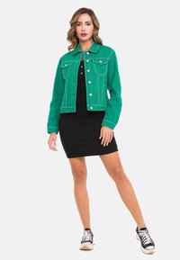 Grüne Jeansjacke mit zwei Brusttaschen, kontrastierenden weißen Nähten und Druckknöpfen, getragen über einem schwarzen Kleid und schwarzen Sneakers.