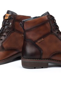 Botas de tobillo de cuero marrón con paneles laterales texturizados, ganchos de cordones negros y tacones de madera apilados, ubicadas sobre un fondo blanco.