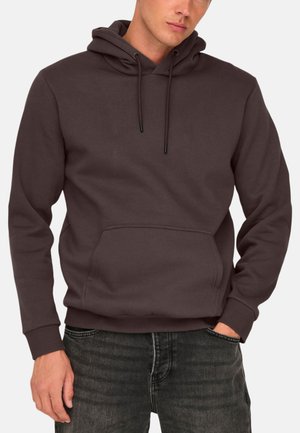 Mann trägt einen dunkelbraunen Hoodie mit Kängurutasche vorne und schwarze Jeans, eine Hand in der Tasche, steht vor einem einfarbigen hellen Hintergrund.