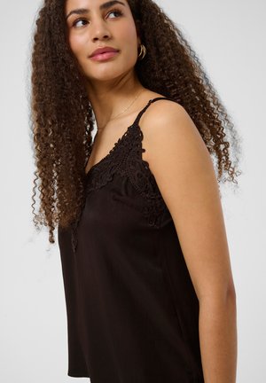 Femme aux longs cheveux bouclés portant un haut sans manches noir bordé de dentelle, regardant sur le côté sur un fond uni.