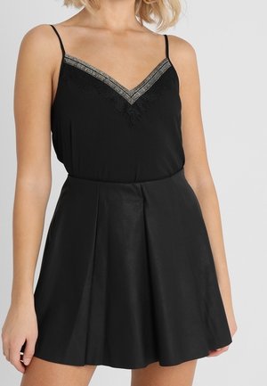 Kobieta ubrana w czarny top bez rękawów z koronkowym wykończeniem oraz czarną spódnicę z wysokim stanem i plisą z przodu.