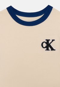 Béžové tričko s krátkým rukávem, navy modrý žebrovaný límec a černé vyšívané logo "cK" na levé straně kapsy na prsou.