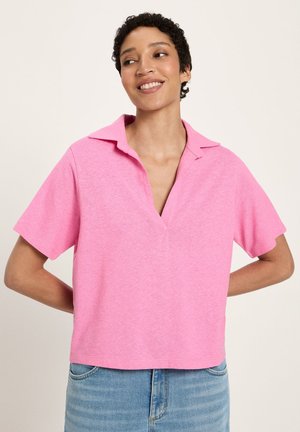 Lächende Person mit kurzen lockigen Haaren, die ein lockeres rosa kurzärmeliges Hemd mit Kragen und hellblaue Jeans vor einem schlichten Hintergrund trägt.