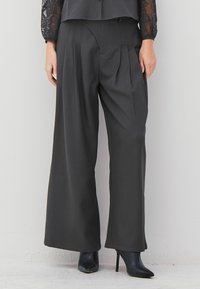 Black wide-leg trousers with front pleats, worn with sheer lace-sleeved top and black pointed high-heeled ankle boots.