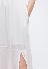 Λευκό φόρεμα με πλαϊνή σχισμή που αποκαλύπτει γόνατο λυγισμένο, με ένα μέρος ενός χεριού που ακουμπά στο ισχίο, πάνω σε απλό φωτεινό φόντο.