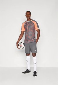 Grå idrettsjersey med oransje detaljer, matchende shorts og hvite sokker, holder en fotball, har merkevarelogo på klærne.