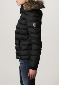 Black puffer jacket featuring a fur-lined hood, horizontal quilted texture, side pockets, and a circular logo patch on the sleeve.